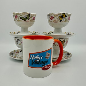 Peut inclure: Une tasse en c&eacute;ramique blanche avec une bordure et une poign&eacute;e orange. La tasse a un autocollant bleu et blanc avec le texte "Holly's Vintage Decor". La tasse est plac&eacute;e devant deux coupelles &agrave; dessert en c&eacute;ramique blanche avec des motifs de papillons.