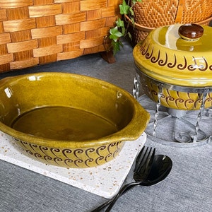 Op de afbeelding: Een ovale, olijfgroene keramische ovenschaal en een bijpassende serveerschaal met deksel. Beide hebben een bruin decoratief patroon. De schaal met deksel staat op een roestvrijstalen standaard. Zwart bestek staat op de voorgrond.