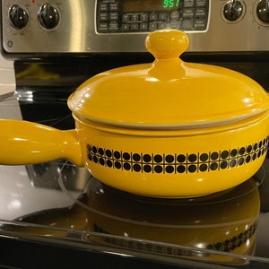 May include: A yellow ceramic pot with a black and white geometric pattern around the rim. The pot has a lid and a handle. The pot is sitting on a black hob.