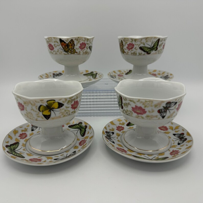 Peut inclure: Ensemble de quatre bols &agrave; dessert en porcelaine blanche avec soucoupes assorties. Chaque bol et soucoupe pr&eacute;sente un motif floral avec des papillons dans des tons de jaune, vert, orange et marron.