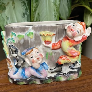 May include: A ceramic planter shaped like a tree trunk with two elves painted on it. The elf on the right is holding a yellow flower and the elf on the left is lying down with their legs crossed. The planter is painted in shades of brown, green, yellow, red, blue, and pink.