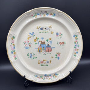 May include: Cream ceramic plate with a rural scene design. The plate has a gold rim and a central illustration of houses, trees, and farm animals. Floral and bird motifs decorate the border.