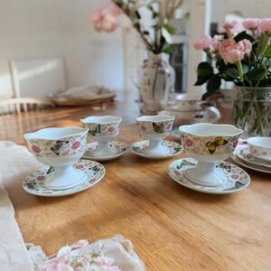 Peut inclure: Quatre coupes &agrave; dessert en porcelaine blanche avec des motifs de papillons et des finitions dor&eacute;es. Chaque coupe est sur une soucoupe assortie avec un bord festonn&eacute;. Les coupes sont dispos&eacute;es sur une table en bois.