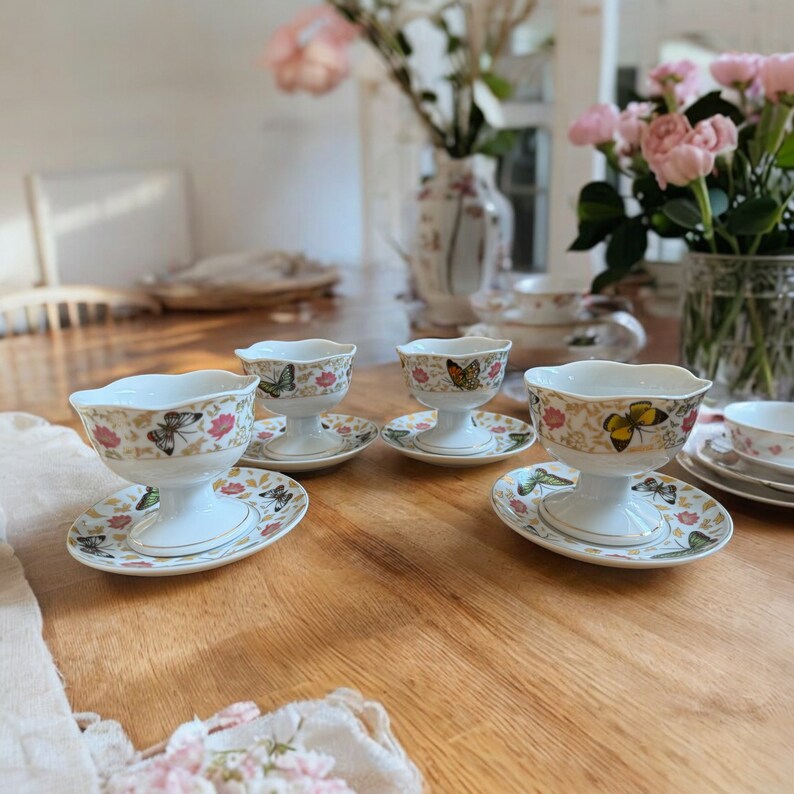 Peut inclure: Quatre coupes &agrave; dessert en porcelaine blanche avec des motifs de papillons et des finitions dor&eacute;es. Chaque coupe est sur une soucoupe assortie avec un bord festonn&eacute;. Les coupes sont dispos&eacute;es sur une table en bois.