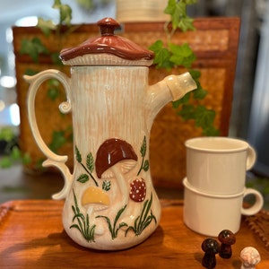 May include: A ceramic teapot shaped like a tree trunk with mushroom decorations. The teapot has a brown lid and a handle. There are three small ceramic mushrooms on a wooden surface.