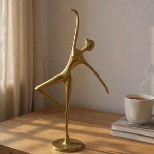May include: A gold-coloured metal sculpture of a dancer in a graceful pose. The figure is slender with one leg extended and arms reaching upwards and outwards. The sculpture is mounted on a circular base, and is displayed on a wooden surface.
