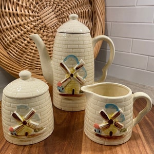 Può includere: Un set da tè in ceramica vintage con un design a mulino a vento. Il set comprende una teiera, un creamer e una zuccheriera. Ogni pezzo è decorato con un motivo a mattoni e un mulino a vento colorato.