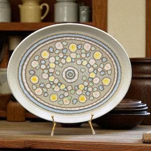 Puede incluir: Un plato de cerámica con un diseño floral. El plato tiene un borde beige y un patrón central de motivos florales amarillos, azules y verdes. El plato se exhibe sobre un soporte dorado, mostrando su atractivo decorativo.