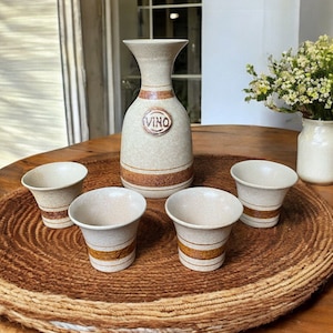 May include: A set of four small, white ceramic cups with brown bands and a larger, white ceramic jug with brown bands and the word "VINO" on the front. The cups and jug are on a brown woven mat.