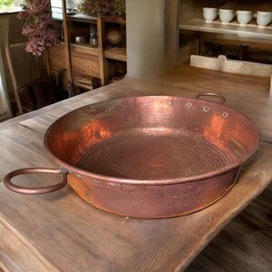 May include: A large, copper, round serving bowl with two handles. The bowl has a hammered texture and a slightly worn patina.