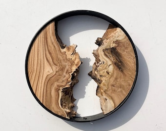 May include: A round wooden wall decor with a natural wood grain pattern. The wood is split in two, creating a unique design. The wood is framed by a black metal ring.