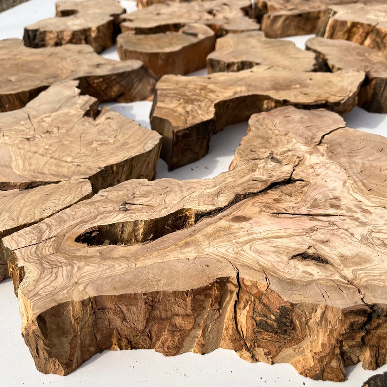 May include: A collection of natural wood slices, each with unique grain patterns and textures. The slices are various sizes and shapes, with some having a rough, bark-like edge. The wood is a light brown color with darker brown streaks.