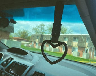 May include: A black heart-shaped car charm hanging from the rearview mirror of a car. The charm is made of a soft, flexible material and has a smooth, shiny finish.