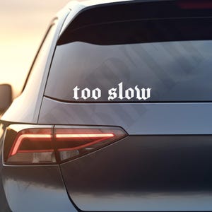 Könnte beinhalten: Ein weißer Vinyl-Aufkleber mit dem Schriftzug "too slow" in gotischer Schrift ist auf der Heckscheibe eines dunkelgrauen Autos angebracht. Das Rücklicht des Autos leuchtet, der Hintergrund ist unscharf.