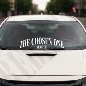 Pode incluir: Carro branco com uma faixa no para-brisa que diz "THE CHOSEN ONE REBIRTH" em letras brancas. O carro está estacionado em uma rua, com árvores ao fundo.