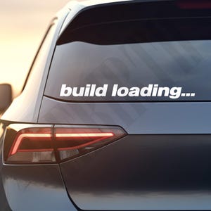 Könnte beinhalten: Ein weißer Aufkleber mit dem Text "build loading..." ist auf der Heckscheibe eines dunkelgrauen Autos angebracht. Das Rücklicht des Autos leuchtet, und der Hintergrund zeigt einen weichen, warmen Sonnenuntergang.