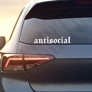 May include: A white vinyl decal with the word "antisocial" in a gothic font is affixed to the rear window of a dark gray car. The car's taillight is illuminated, and the background shows a soft, blurred sunset.