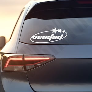 Puede incluir: Adhesivo ovalado blanco con la palabra "wasted" en fuente estilizada y tres estrellas. Se encuentra en la ventana trasera de un coche gris oscuro, con la luz trasera visible.