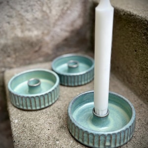 Può includere: Tre portacandele in ceramica color verde acqua con un design a coste. Un portacandele ha una candela bianca inserita. I portacandele sono su una superficie di pietra grigia. I portacandele hanno un design circolare con un inserto metallico per tenere la candela.