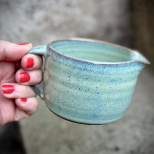 Op de afbeelding: Handgemaakte keramische kan met een handvat, met een lichtblauwe en groene glazuur. De kan heeft een tuit om gemakkelijk te schenken en een rustieke, getextureerde uitstraling. Het aardewerk is ideaal voor het serveren van sauzen of dressings.