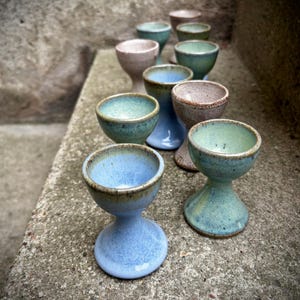 Könnte beinhalten: Eine Sammlung handgefertigter Keramik-Eierbecher in verschiedenen Farben, darunter Blau, Grün und Beige. Jeder Becher hat eine einzigartige Form und Glasur, mit einem Sockel und einer kleinen Schale. Die Becher sind auf einer Betonoberfläche angeordnet.