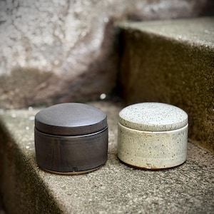 Könnte beinhalten: Zwei Keramikdosen mit Deckel. Eine Dose ist dunkelgrau, die andere gesprenkelt cremefarben. Beide sind zylindrisch mit abgerundeten Deckeln. Die Dosen stehen auf einer Betonoberfläche.