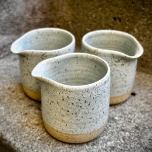 Può includere: Tre brocche per panna in ceramica fatte a mano. Le brocche sono azzurre con dettagli maculati e una base beige naturale. Ogni brocca ha un beccuccio per versare facilmente. Le brocche sono disposte su una superficie strutturata.