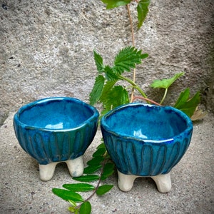 May include: Two small blue ceramic bowls with white feet. The bowls have a textured, ribbed design and are sitting on a grey concrete surface.