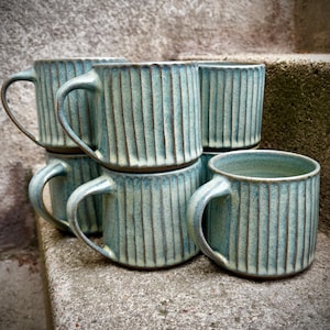 May include: Six teal ceramic mugs with a ribbed design, each with a curved handle. The mugs are stacked on a concrete surface. The mugs have a cylindrical shape.