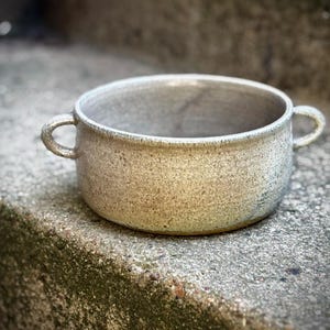 Puede incluir: Un cuenco de cerámica moteada con dos asas pequeñas. El cuenco tiene una forma ancha y poco profunda y una paleta de colores neutros con toques de beige y gris. La superficie texturizada añade una sensación rústica y hecha a mano. Ideal para servir sopas, guisos u otros platos.