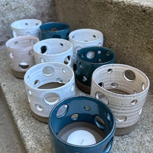 May include: A collection of ceramic candle holders in various colors, including white, teal, and beige. Each cylindrical holder features multiple circular cutouts and a brown base. A lit tealight candle is visible in one holder.