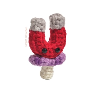 May include: A crocheted magnet toy shaped like a horseshoe with a purple and white mushroom base. The magnet is red with gray ends and has black eyes.