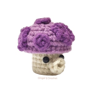May include: A handmade crochet mushroom in shades of purple and cream. The mushroom cap is a gradient of purple hues, with decorative spiral accents. The stem is cream-colored, and it has black button eyes and a small, rounded mouth. The text "Ginger | Crochet" is at the bottom.