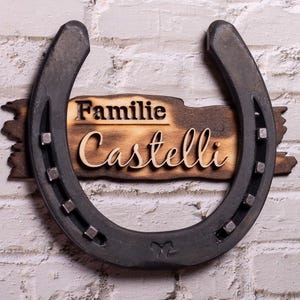K&ouml;nnte beinhalten: Ein Holzschild mit dem Text "Familie Castelli" auf einem schwarzen Hufeisen. Das Schild ist abgenutzt und hat einen rustikalen Look.