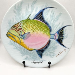 May include: A decorative plate featuring a colorful triggerfish design. The fish has a pink belly, yellow and green body, and blue fins. The plate has a light blue background with a coral-like pattern and the word "Triggerfish" written on it.