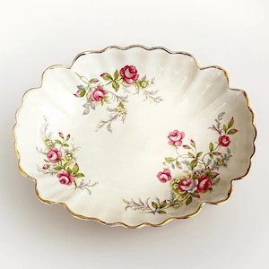 May include: An oval-shaped ceramic dish with a scalloped edge and a gold-painted rim. The white surface features delicate floral illustrations of pink roses with green leaves and small yellow accents arranged in clusters around the interior.