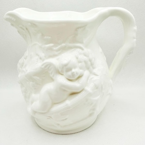 May include: White ceramic pitcher with a detailed relief design. The pitcher features a cherub and floral motifs. It has a curved handle and a scalloped rim. The pitcher is a decorative item.