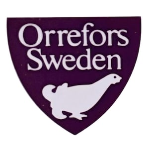 Op de afbeelding: Een paars schildvormig embleem met de woorden "Orrefors Sweden" in het wit. Een witte vogelafbeelding staat onderaan. Het embleem is waarschijnlijk een logo of een badge.