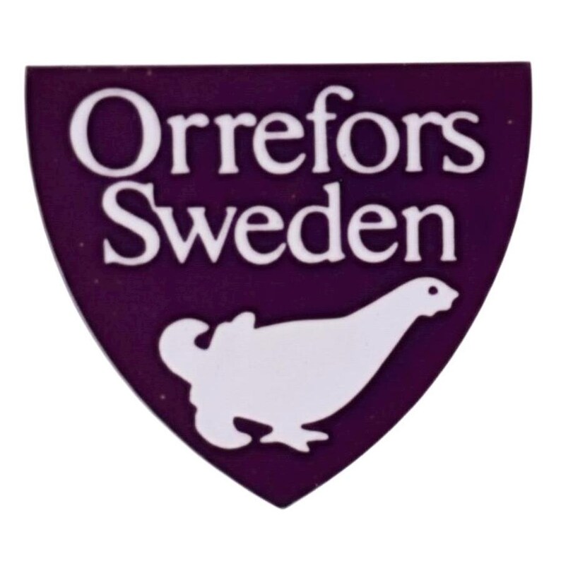 Op de afbeelding: Een paars schildvormig embleem met de woorden "Orrefors Sweden" in het wit. Een witte vogelafbeelding staat onderaan. Het embleem is waarschijnlijk een logo of een badge.