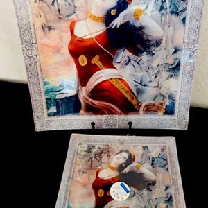 May include: Two decorative glass plates featuring a painting of a woman in a red dress with gold accents. The plates have a rectangular shape with a decorative border. The artwork has a vintage aesthetic.