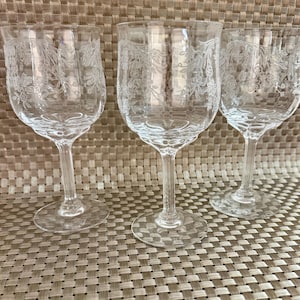 May include: Three clear wine glasses with etched floral designs. The glasses have a rounded bowl shape, slender stems, and circular bases. The etched patterns are visible throughout the bowl of each glass. The glasses are arranged on a textured surface.