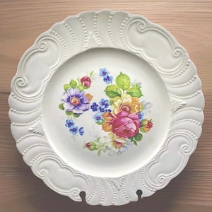 May include: White decorative plate with a floral design. The plate features a raised, ornate border and a central design of colorful flowers, including purple, pink, yellow, and blue blooms with green leaves. The plate has a vintage aesthetic.