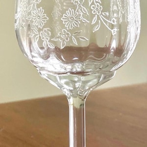 May include: A clear, stemmed wine glass with a floral etched design. The glass features a rounded bowl, a slender stem, and a circular base. The etched pattern includes flowers and leaves, adding a delicate touch to the glassware.