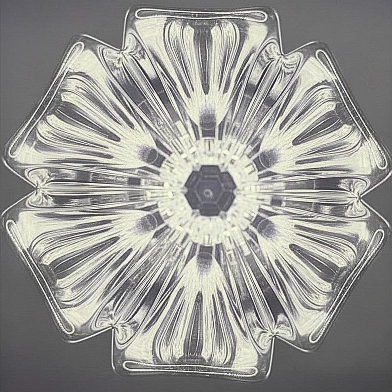 Op de afbeelding: Een helder, glazen decoratief object met een bloemachtig ontwerp. Het object heeft zes bloemblad-vormige secties die uitstralen vanuit een centrale zeshoekige kern. Het glas heeft een getextureerd, geribbeld patroon, waardoor een spel van licht en schaduw ontstaat. De algehele vorm is symmetrisch en elegant.