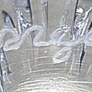 Op de afbeelding: Close-up van een helder glazen object met een getextureerd oppervlak. Het woord "Serena" is in cursief schrift over het midden van het glas geschreven. Het glas heeft een reflecterende kwaliteit.