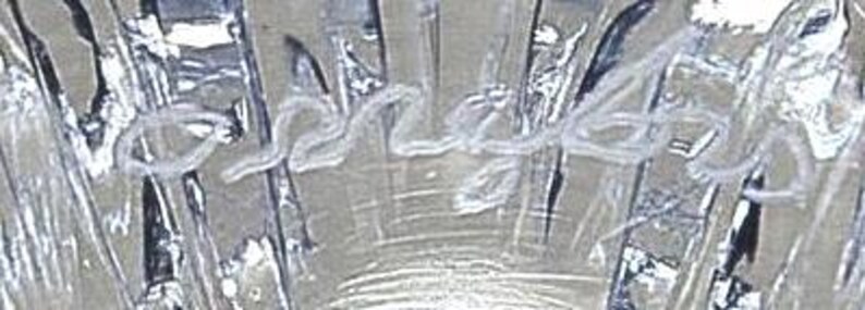Op de afbeelding: Close-up van een helder glazen object met een getextureerd oppervlak. Het woord "Serena" is in cursief schrift over het midden van het glas geschreven. Het glas heeft een reflecterende kwaliteit.