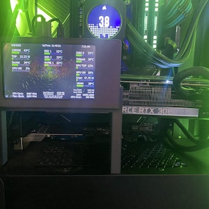 May include: A close-up of a computer's internal components, including a graphics card with the text "FORCE RTX 3080" and a monitor displaying system information. The monitor shows the CPU temperature, GPU temperature, and other system statistics. The computer is lit with green LED lights.