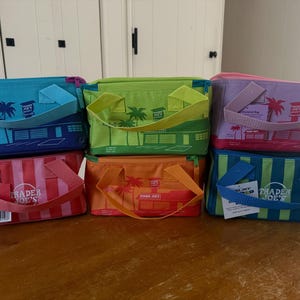 Pode incluir: Uma coleção de sacolas térmicas coloridas. Cada sacola apresenta um design de palmeira e edifício, com as palavras "Trader Joe's" impressas na frente. As sacolas vêm em várias cores, incluindo azul, verde, rosa, laranja e vermelho.
