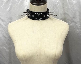 Könnte beinhalten: Schwarze Lederhalskette mit silbernen Stacheln und einem herzförmigen Akzent. Die Halskette wird auf einer Schaufensterpuppe präsentiert. Es ist ein Gothic-Accessoire.