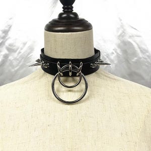 Könnte beinhalten: Schwarze Leder-Halskette mit silberfarbenen Stacheln und zwei großen Metallringen. Die Halskette wird auf einer Schaufensterpuppe präsentiert. Die Stacheln sind gleichmäßig um den Kragen verteilt.